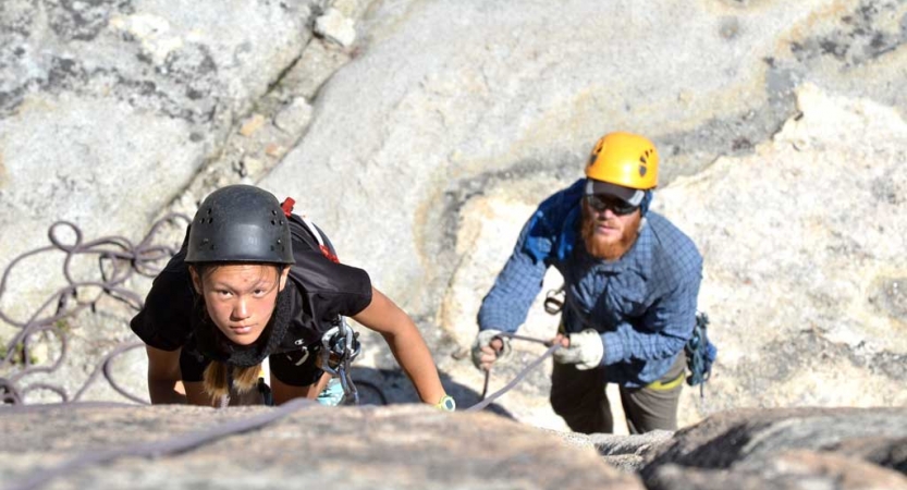A student wearing safety gear looks up at the camera while they climb a rock wall. An instructor is below, belaying them.